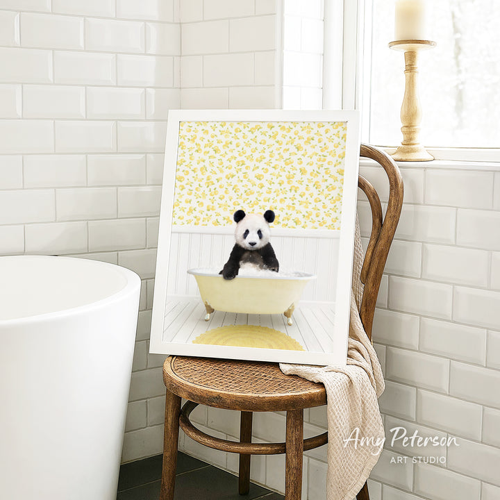 A framed image of a panda sitting in a bathtub, surrounded by yellow flowers, is displayed on a wooden chair in a bathroom setting.