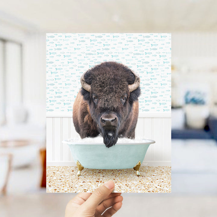 A hand holds up a framed image of a bison in a bathtub, with a patterned wall behind it.