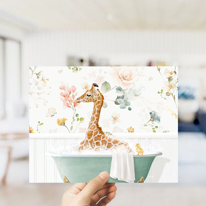 A hand holding a framed print featuring a giraffe sitting in a bathtub filled with water and surrounded by flowers.