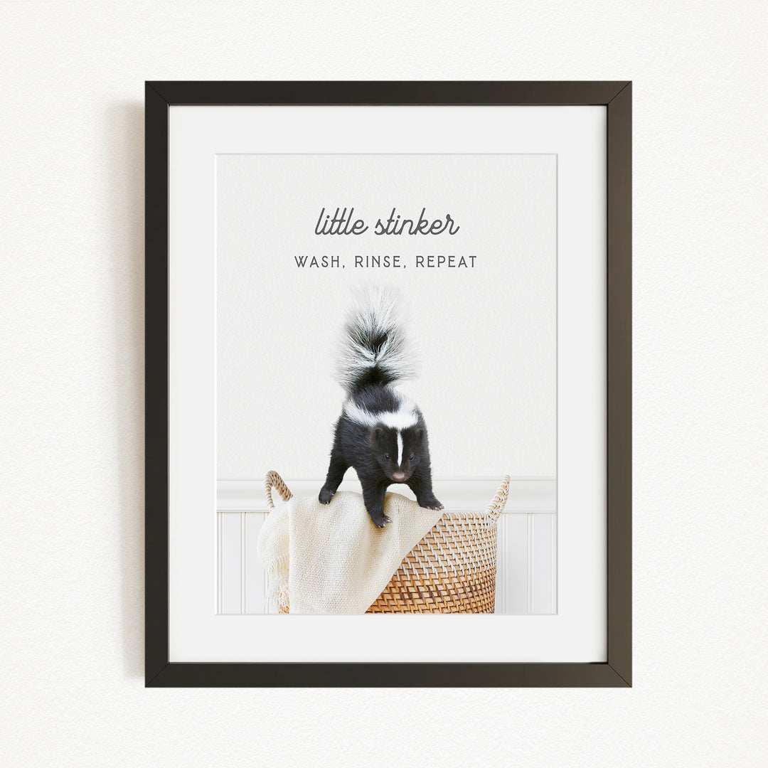 A framed image of a black and white skunk standing on a woven basket, with the text "little stinker" and "WASH, RINSE, REPEAT" written above it.