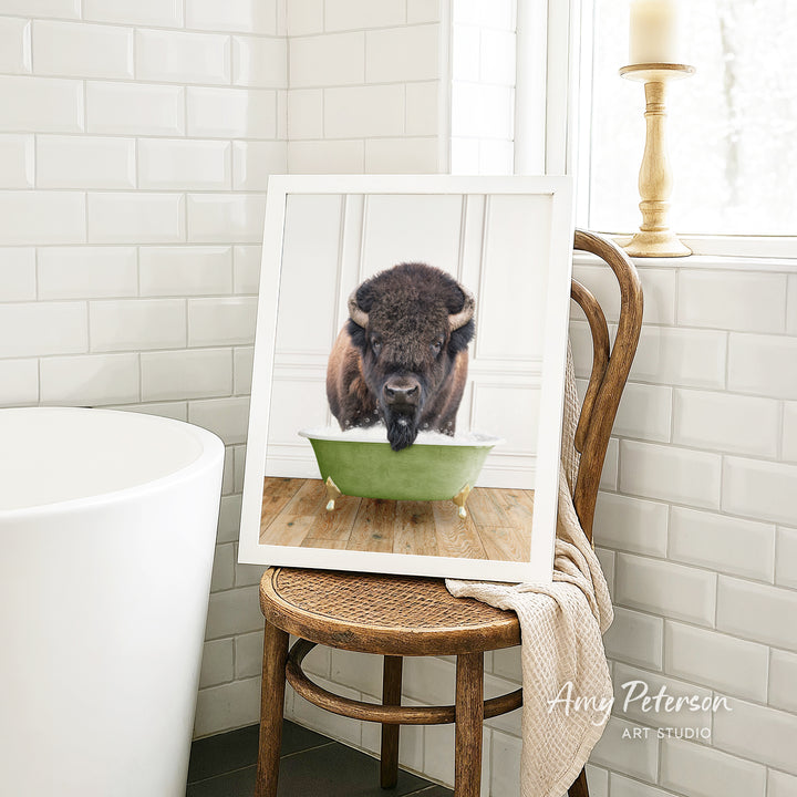 A framed image of a bison sitting in a green bathtub, with a towel draped over the back of the chair.