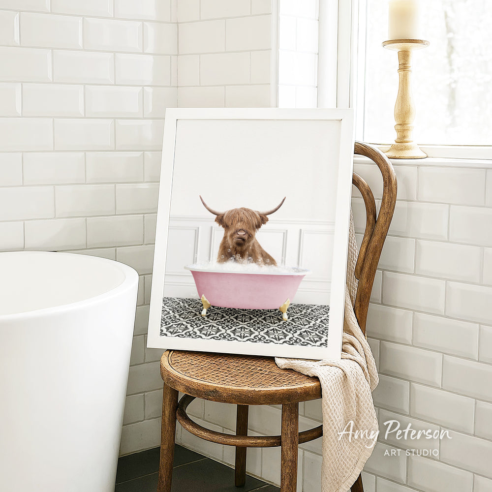 A framed image of a Highland cow sitting in a pink bathtub, with a towel draped over the back of a wooden chair in a bathroom setting.