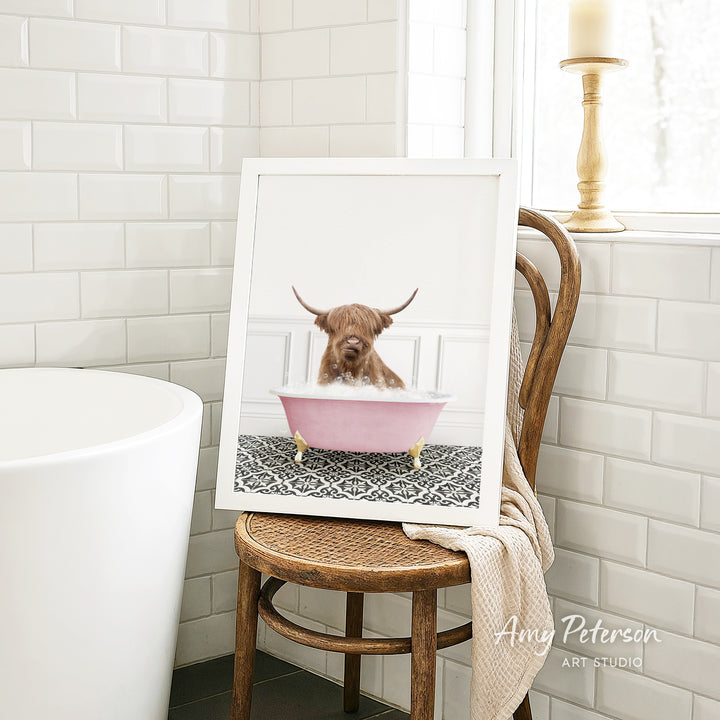 A framed image of a Highland cow sitting in a pink bathtub, with a towel draped over the back of a wooden chair in a bathroom setting.