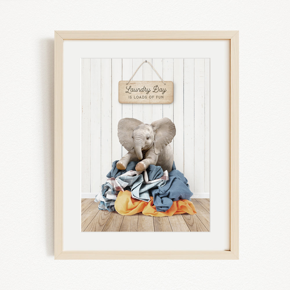 A framed image of a baby elephant sitting on a pile of laundry, with a sign above that says "Laundry Day is loads of fun".