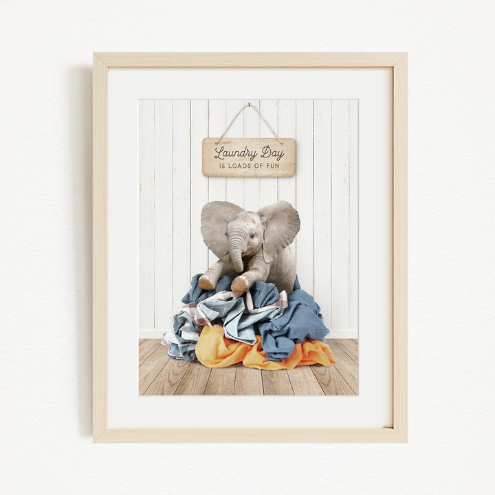 A framed image of a baby elephant sitting on a pile of laundry, with a sign above that says "Laundry Day is loads of fun".