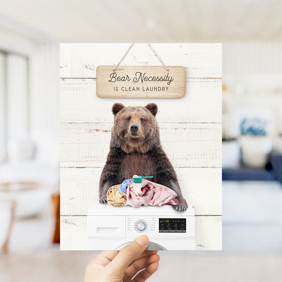 A hand holding a sign with a bear sitting on a washing machine, with a wooden sign above that says "Bear Necessity is Clean Laundry".