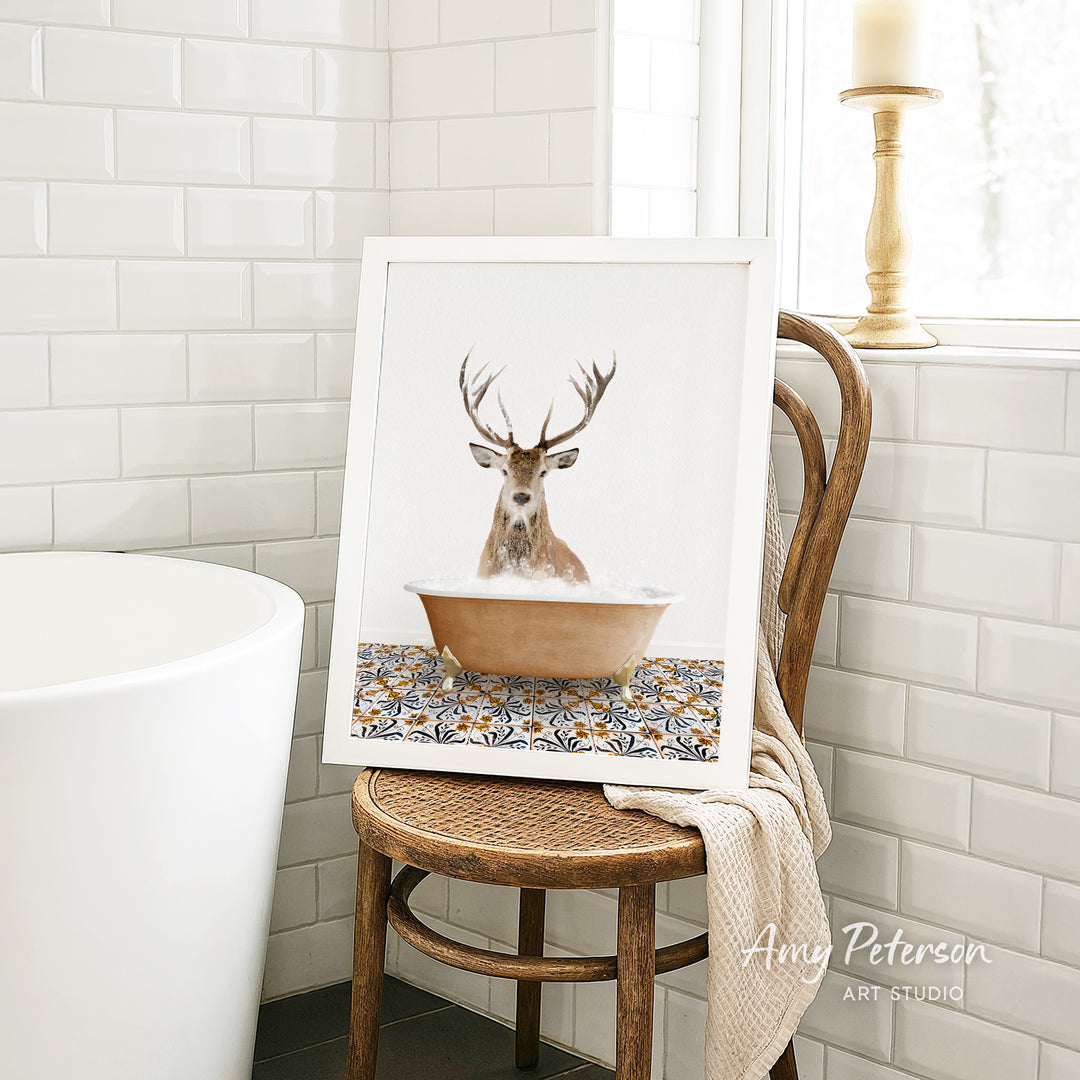 A framed image of a deer sitting in a bathtub, surrounded by a patterned tile design, is displayed on a wooden chair in a bathroom setting.