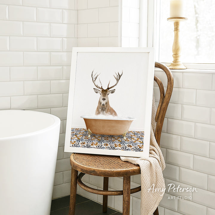 A framed image of a deer sitting in a bathtub, surrounded by a patterned tile design, is displayed on a wooden chair in a bathroom setting.
