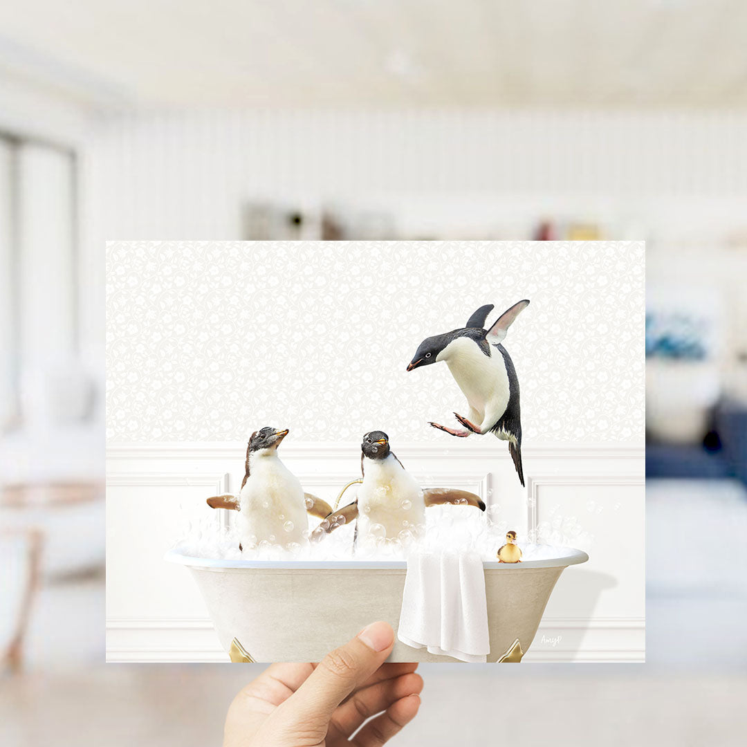 A hand holds up a framed image depicting three penguins sitting in a bathtub, with one penguin jumping out of the tub.