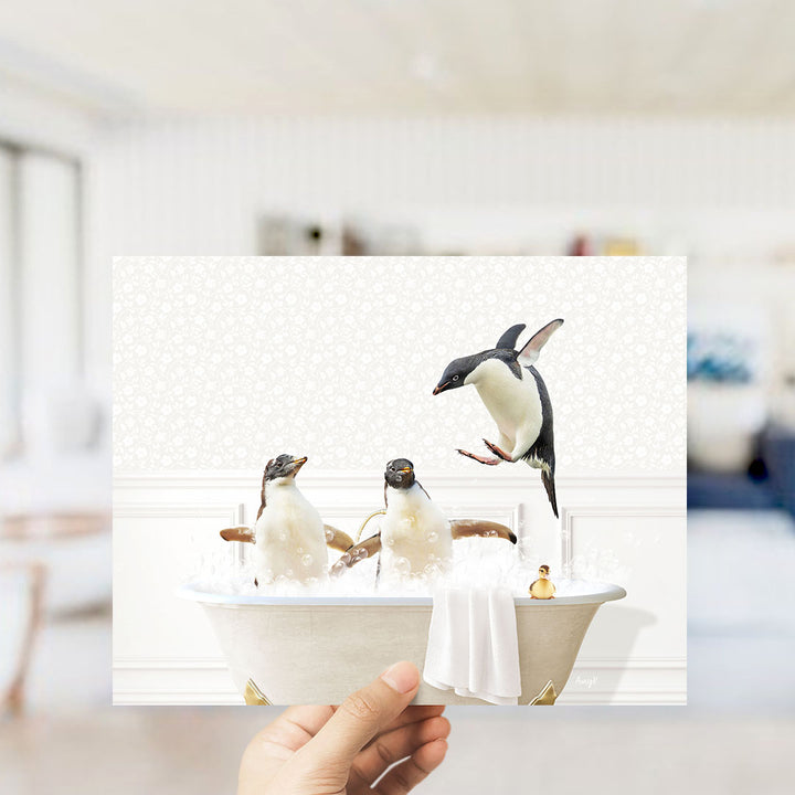 A hand holds up a framed image depicting three penguins sitting in a bathtub, with one penguin jumping out of the tub.