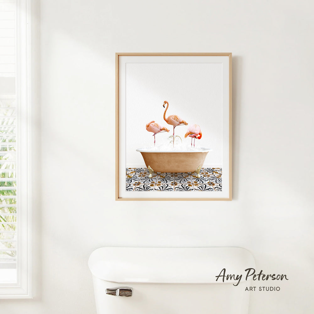 A framed print of three flamingos standing in a bathtub is hanging on a white wall.