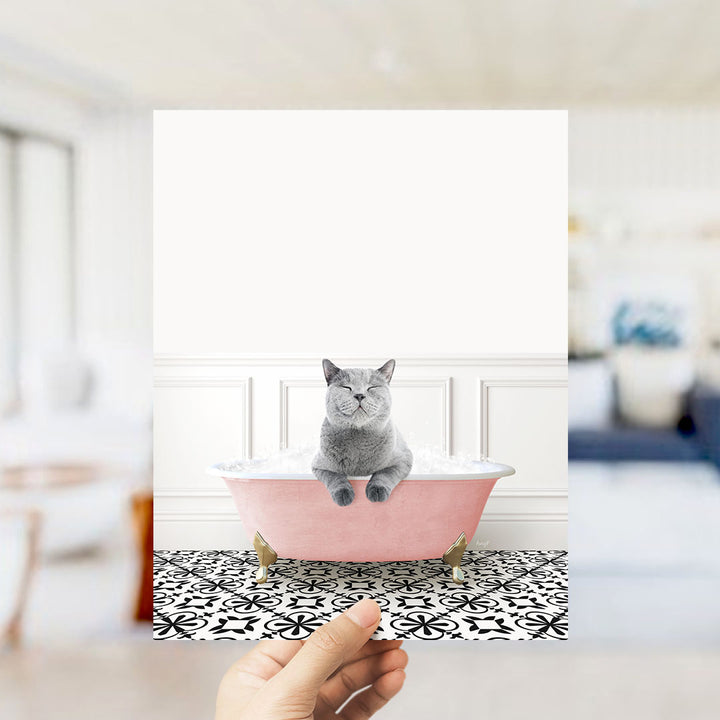 A gray cat sitting in a pink bathtub filled with bubbles, with a white wall and black and white patterned floor in the background.