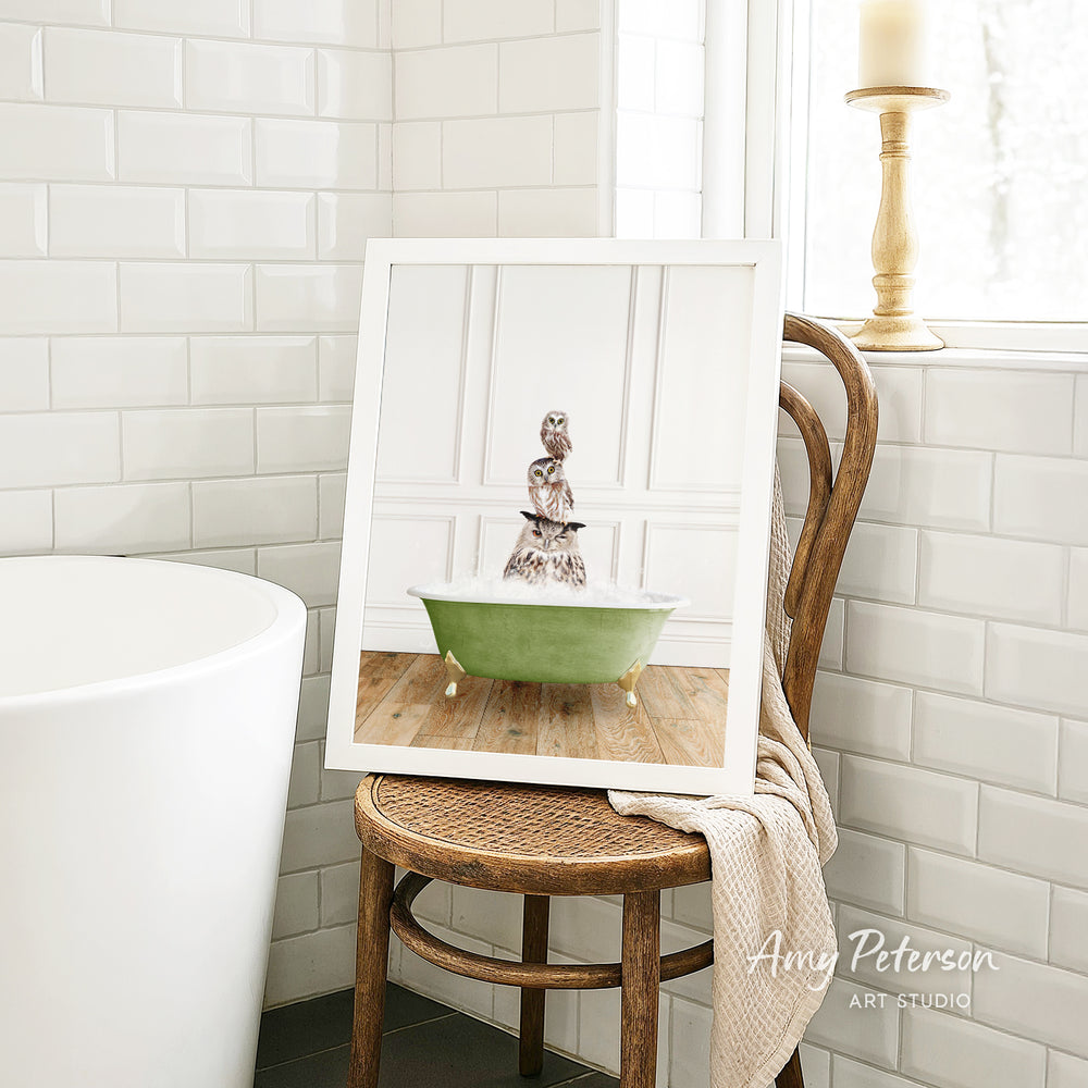 A framed image of a cat sitting in a green bathtub, with a towel draped over the back of a wooden chair in a bathroom setting.