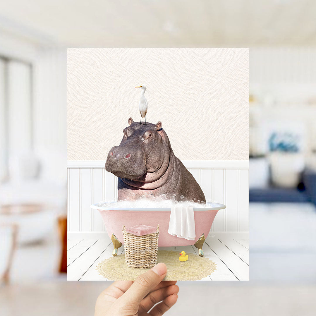 A hand holds up a framed image depicting a hippopotamus sitting in a pink bathtub, with a bird perched on its head.