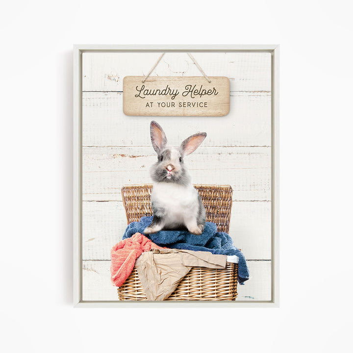 A gray and white rabbit sitting in a wicker laundry basket filled with clothes, with a wooden sign above that reads "Laundry Helper at your service".