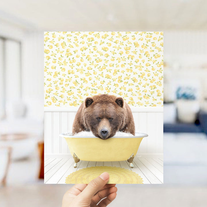 A hand holds up a framed image of a brown bear sitting in a yellow bathtub filled with bubbles, set against a yellow and white floral wallpaper.