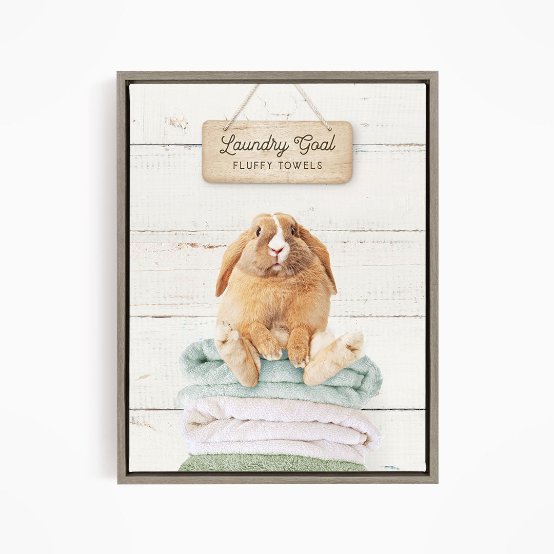 A wooden sign with the text "Laundry Goal Fluffy Towels" hangs above a stack of folded towels, with a rabbit sitting on top of the towels.