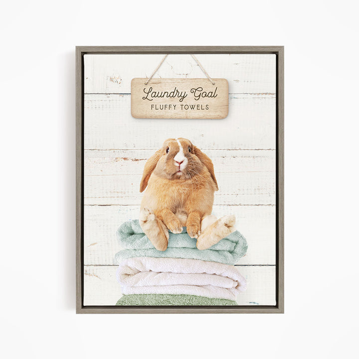 A wooden sign with the text "Laundry Goal Fluffy Towels" hangs above a stack of folded towels, with a rabbit sitting on top of the towels.
