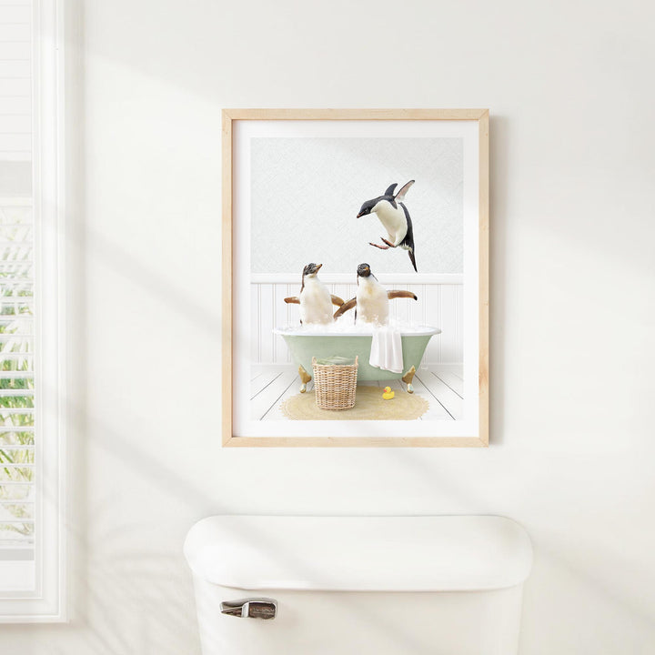 A framed image of two penguins sitting in a bathtub, with one penguin flying towards the other, is hanging on a white wall above a white toilet.