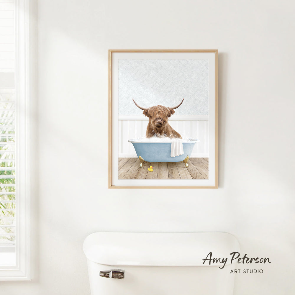 A framed image of a Highland cow sitting in a bathtub, with a towel draped over its back, is hanging on a white wall.