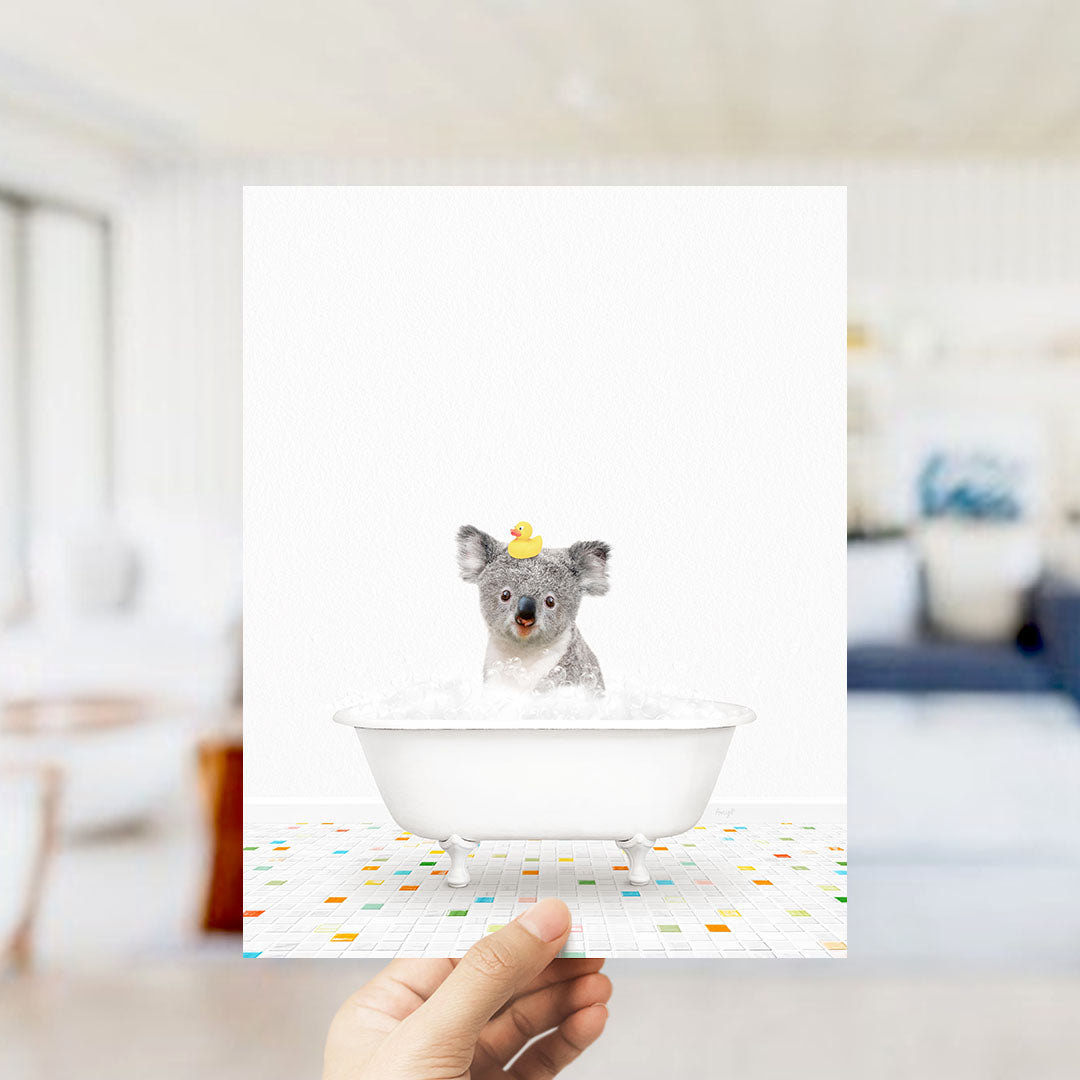 A small gray koala is sitting in a white bathtub filled with bubbles, with a yellow rubber duck floating on its head.