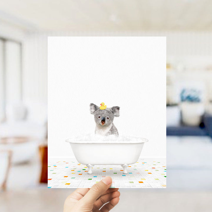 A small gray koala is sitting in a white bathtub filled with bubbles, with a yellow rubber duck floating on its head.