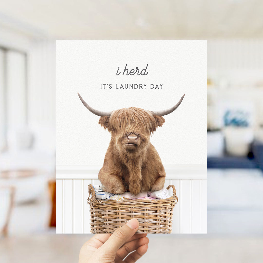 A hand holding a card with a picture of a Highland cow sitting in a wicker basket, with the text "I herd IT'S Laundry Day" printed on it.