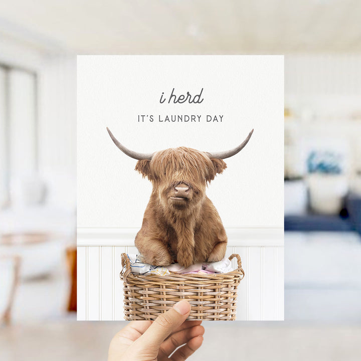 A hand holding a card with a picture of a Highland cow sitting in a wicker basket, with the text "I herd IT'S Laundry Day" printed on it.