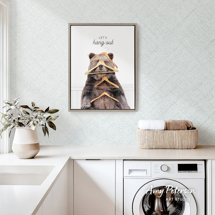 a framed poster of a bear holding a bunch of orange objects, which appears to be a creative and whimsical artwork. The poster is displayed on a wall above a white countertop, which has a basket and a plant on it. The overall setting seems to be a kitchen or a laundry room, as there is a washing machine visible in the background.
