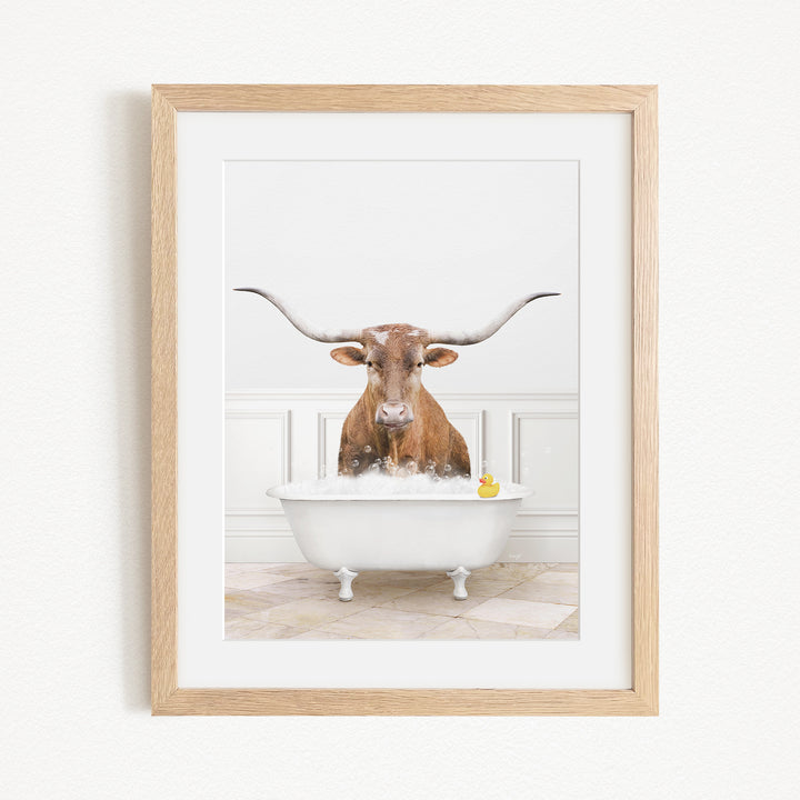 A brown cow with long horns is sitting in a white bathtub filled with bubbles, with a yellow rubber duck floating in the water.