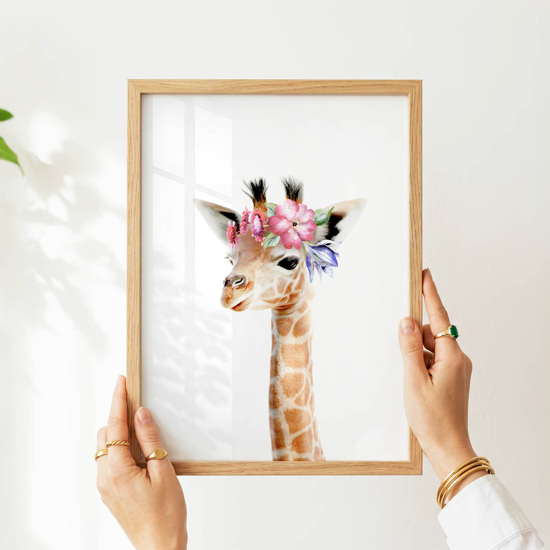 A person's hands are holding a wooden frame with a framed image of a giraffe wearing a flower crown.