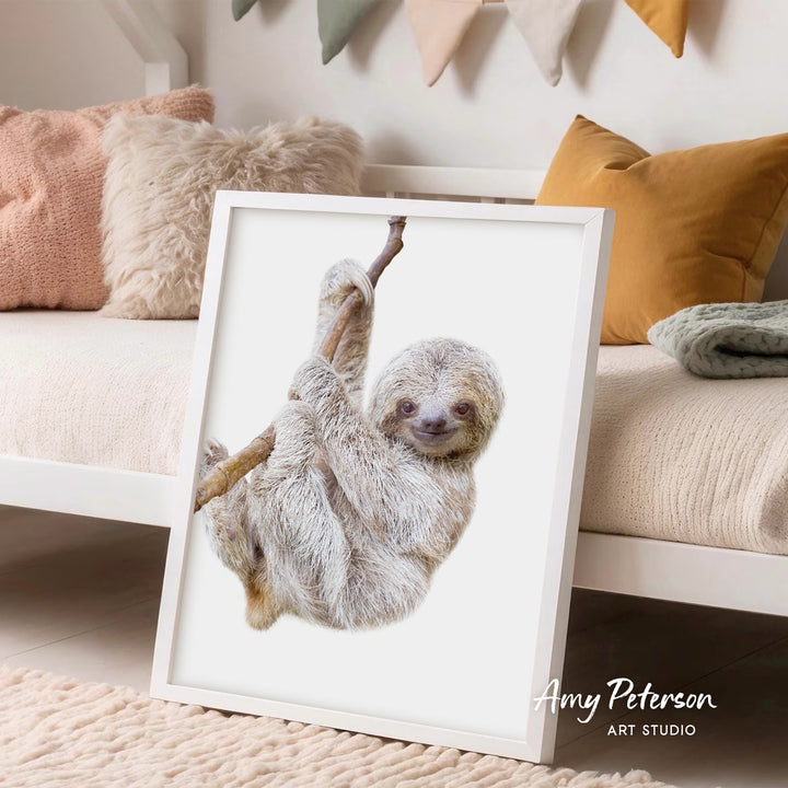 A framed image of a sloth hanging from a branch, displayed in a room with a white bed and colorful pillows.