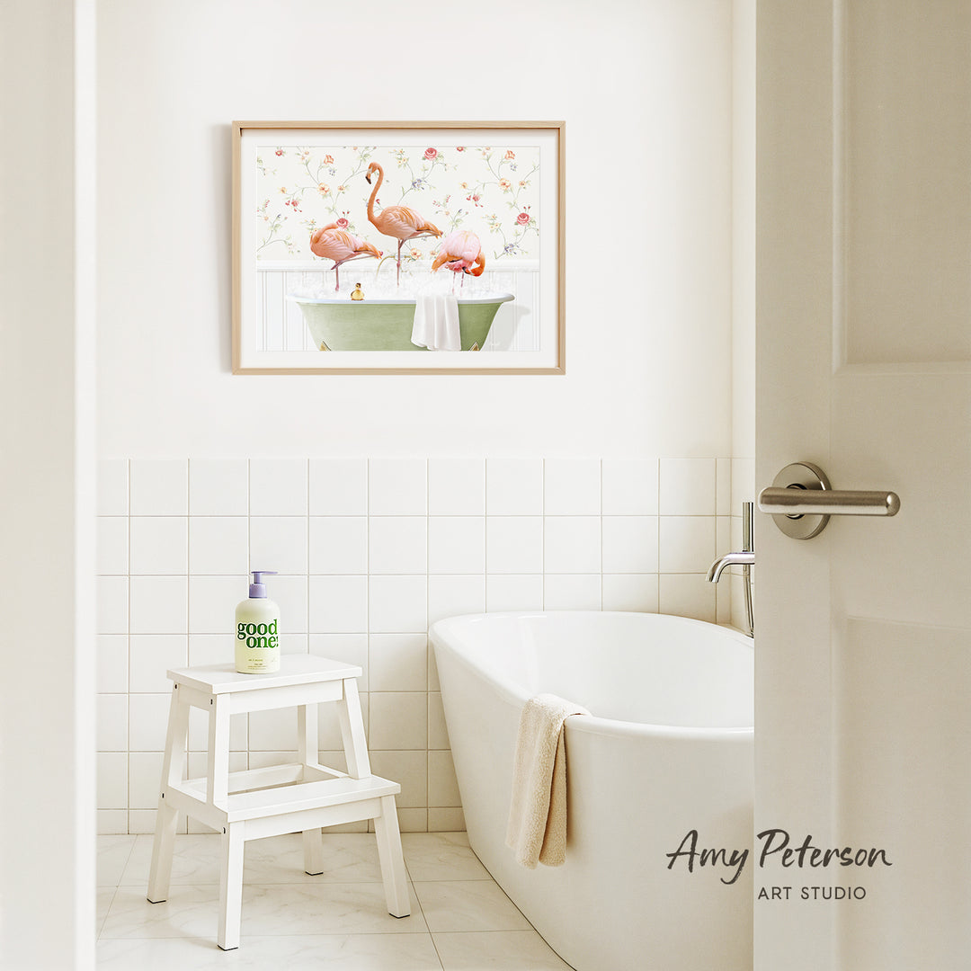 A framed painting of two pink flamingos sitting in a green bathtub is hanging on a white tiled wall above a white bathtub in a bathroom.