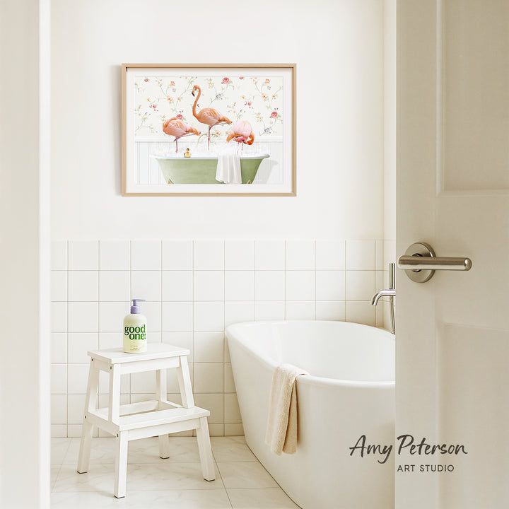 A framed painting of two pink flamingos sitting in a green bathtub is hanging on a white tiled wall above a white bathtub in a bathroom.