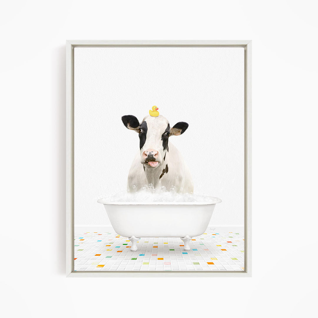 A black and white cow is sitting in a white bathtub filled with bubbles, with a yellow rubber duck perched on its head.