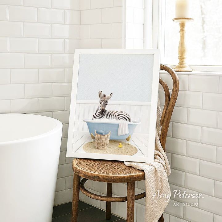 A framed image of a zebra sitting in a bathtub, surrounded by a basket of toys, is displayed on a wooden chair in a bathroom setting.