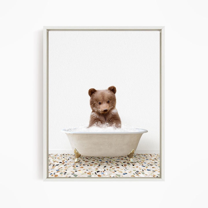A cute brown bear cub sitting in a white bathtub filled with water, looking directly at the camera.
