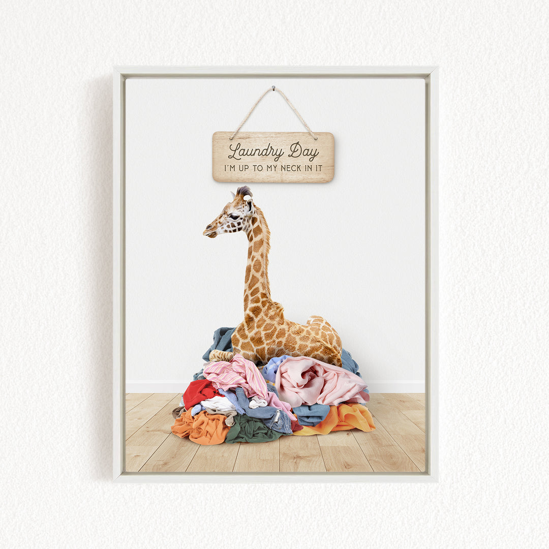 A giraffe standing on a pile of clothes, with a wooden sign above that says "Laundry Day I'm up to my neck in it".