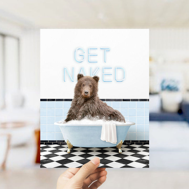 A hand holding a printed image of a brown bear sitting in a bathtub with the text "GET NAKED" displayed above it.