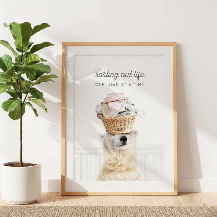 A polar bear balancing a basket of laundry on its head in a framed artwork, with the text "sorting out life one load at a time" displayed above the bear.
