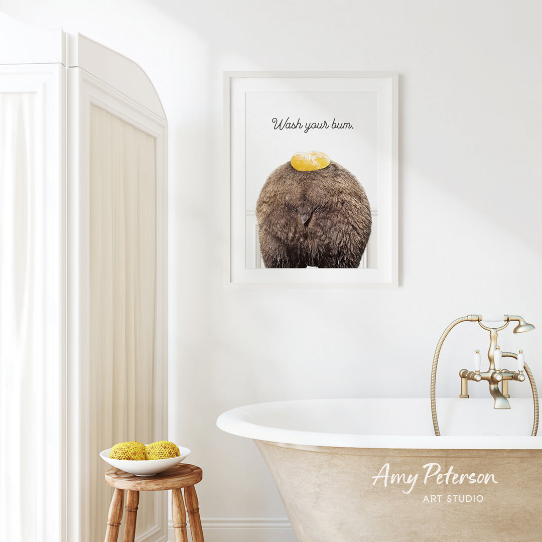 a bathroom with a framed poster of a brown animal, a wooden stool, a bowl of fruit, and a bathtub with a faucet.