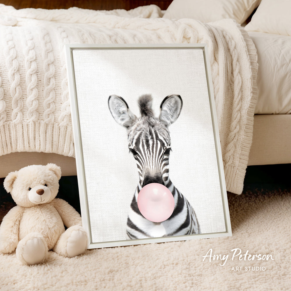 A framed print of a zebra blowing a bubble gum bubble is displayed on a beige carpet, accompanied by a teddy bear.