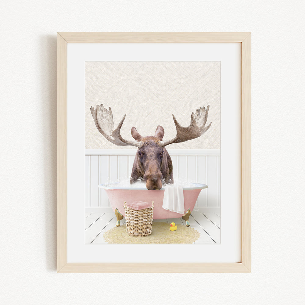 A moose with large antlers is sitting in a pink bathtub, surrounded by a yellow rubber duck and a basket.