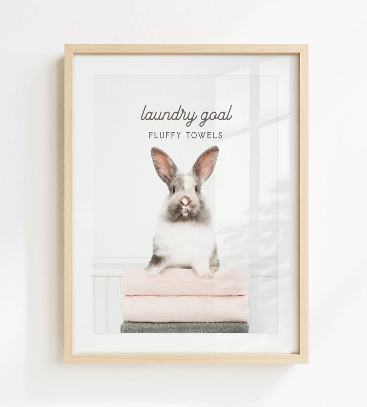 A framed image of a fluffy white rabbit sitting on a stack of pink towels, with the text "laundry goal" and "FLUFFY TOWELS" displayed above the rabbit.