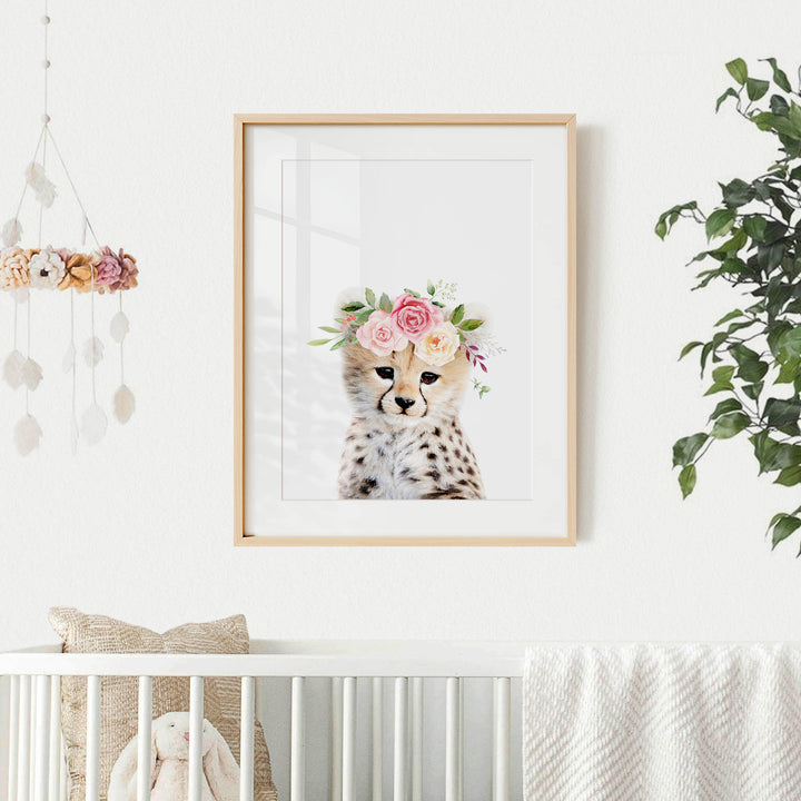 A framed print of a cheetah wearing a floral crown is displayed on a white wall, with a crib and a hanging mobile with flowers in the background.