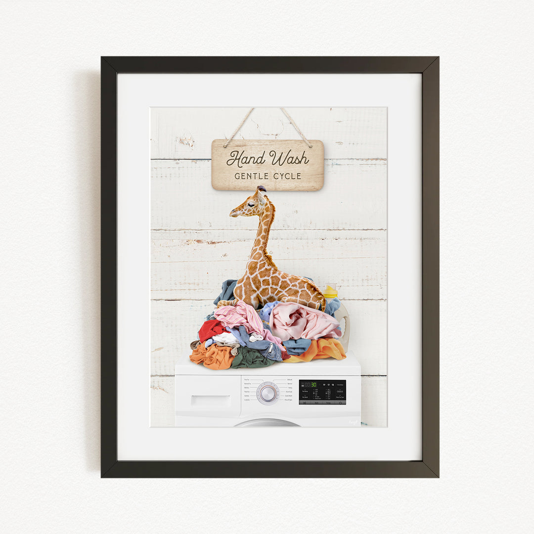 A framed image depicting a giraffe sitting atop a pile of laundry, with a sign above that reads "Hand Wash Gentle Cycle".