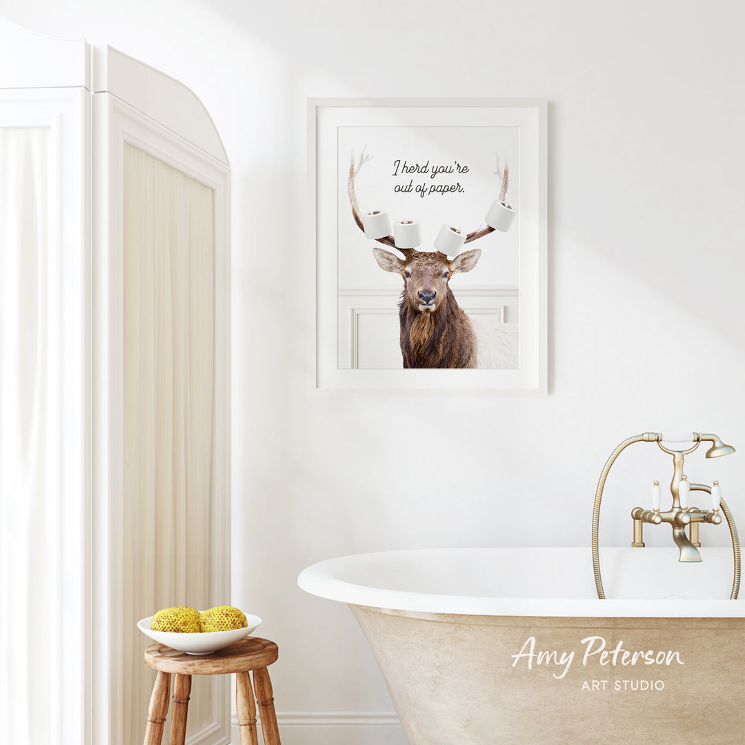 a bathroom with a framed picture of a deer with antlers, a wooden stool, a bowl of fruit, and a bathtub with a faucet.