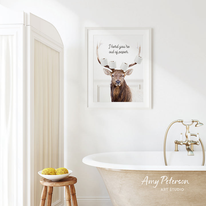 a bathroom with a framed picture of a deer with antlers, a wooden stool, a bowl of fruit, and a bathtub with a faucet.