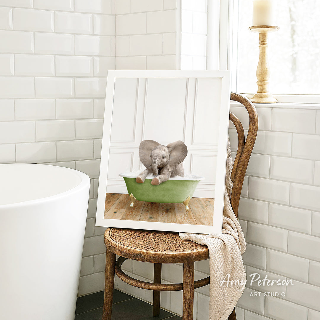 A framed image of a baby elephant sitting in a green bathtub, with a towel draped over the back of a wooden chair in a bathroom setting.