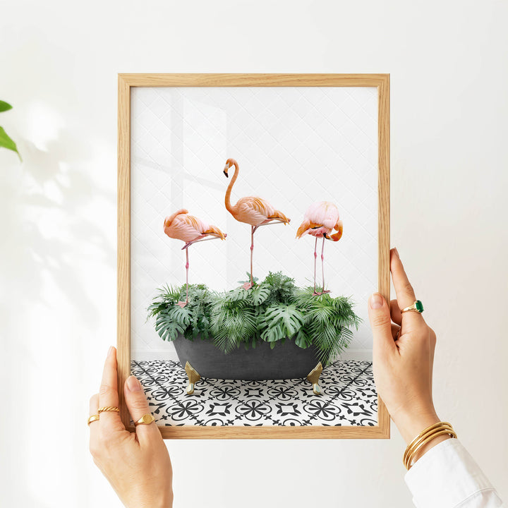 A person's hands are holding a framed picture of three pink flamingos standing on a green plant in a black pot, with a black and white patterned tile floor in the background.